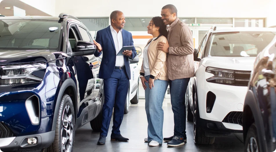 Casal negro sorridente com revendedor de carros no showroom, olhando para o novo SUV