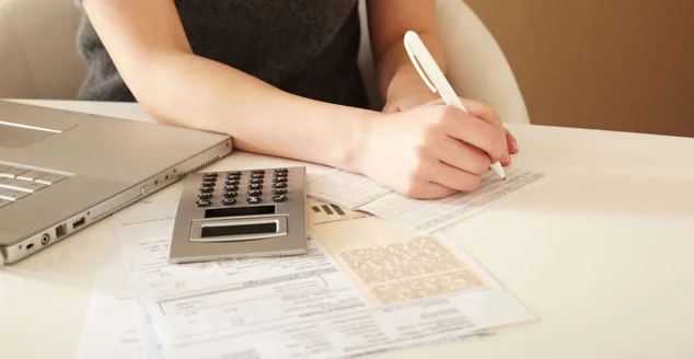 papeis, notebook e calculadora na mesa, enquanto escreve no papel