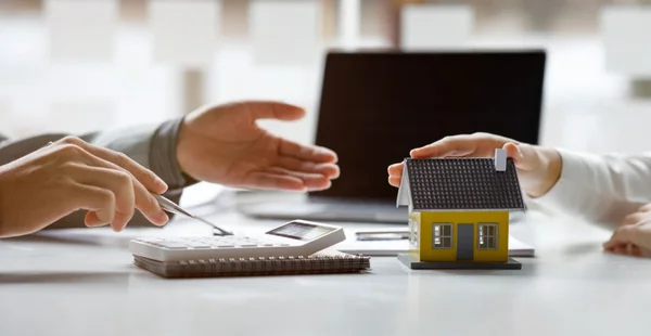 Duas mãos: uma segurando uma caneta e a outra gesticulando. Uma mesa com uma calculadora e um modelo em miniatura de uma casa. Ao fundo, um laptop aberto. Ambiente de escritório, possivelmente relacionado a finanças ou planejamento imobiliário.