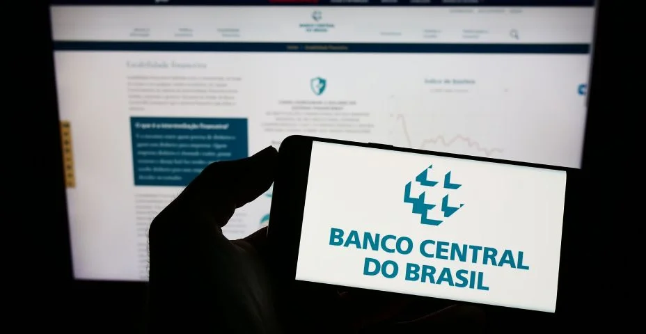 Pessoa segurando celular com logotipo do Banco Central do Brasil (BCB) na tela em frente à página da web. Foco na tela do telefone.