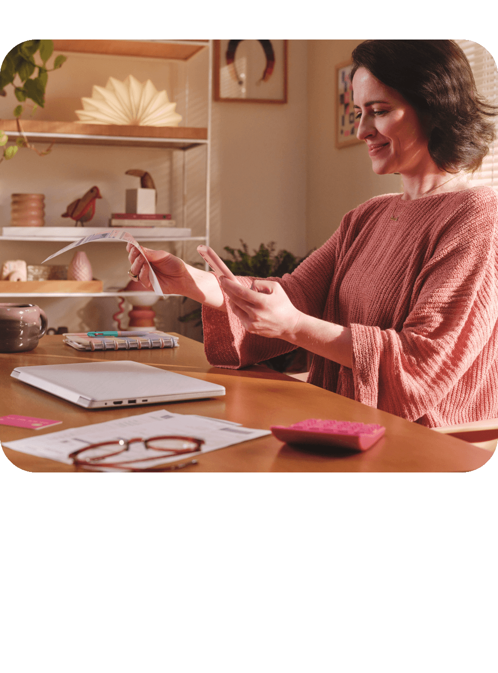 Mulher sorridente usando celular em casa, sentada à mesa com notebook e xícara ao lado.