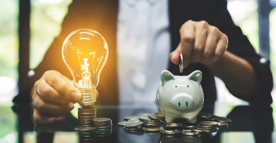 Uma empresária colocando moeda em cofrinho e uma lâmpada sobre pilha de moedas sobre a mesa