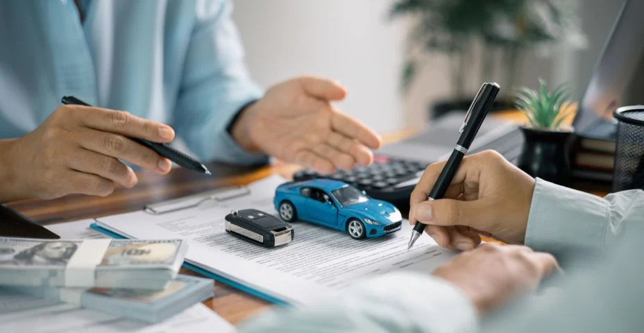 Duas pessoas sentadas à mesa discutindo a compra ou financiamento de um carro. Uma delas aponta para documentos enquanto a outra assina. Na mesa há pilhas de dinheiro, um carrinho de brinquedo azul, uma chave de carro, uma calculadora e materiais de escritório.