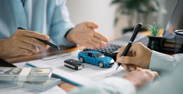Duas pessoas sentadas à mesa discutindo a compra ou financiamento de um carro. Uma delas aponta para documentos enquanto a outra assina. Na mesa há pilhas de dinheiro, um carrinho de brinquedo azul, uma chave de carro, uma calculadora e materiais de escritório.