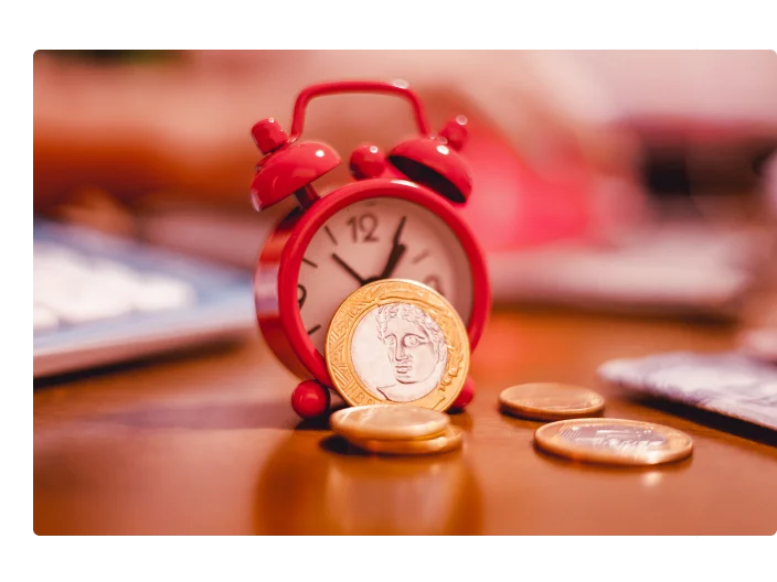 Moedas brasileiras e um despertador vermelho que estão em uma mesa de madeira.