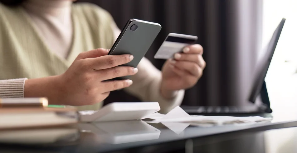 Jovem segurando o celular e o cartão de crédito, sentado à mesa. Mulher feliz faz compras on-line usando pagamento móvel. serviço de aplicativo bancário