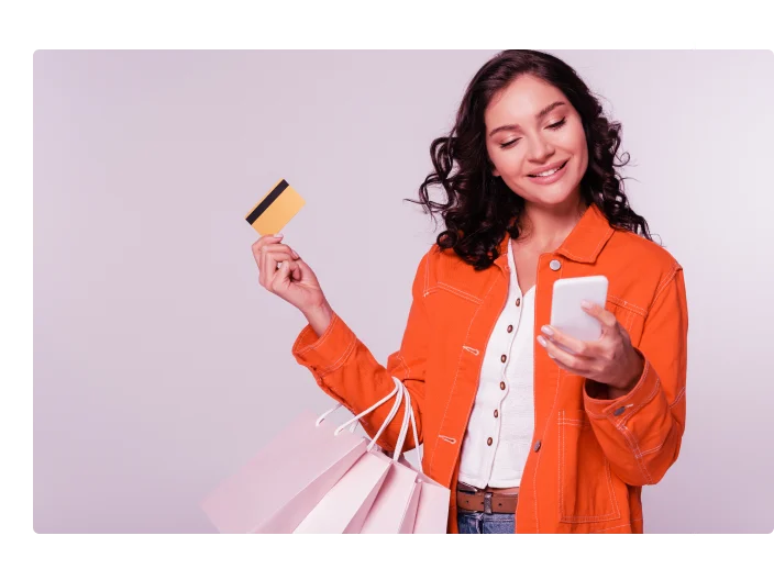 mulher segurando um cartão de crédito na mão esquerda e o celular na mão direita, com sacolas de compra no braço