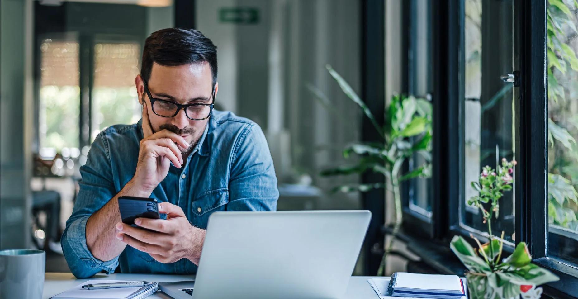 Homem adulto ocupado, comparando notas em seu telefone com laptop.