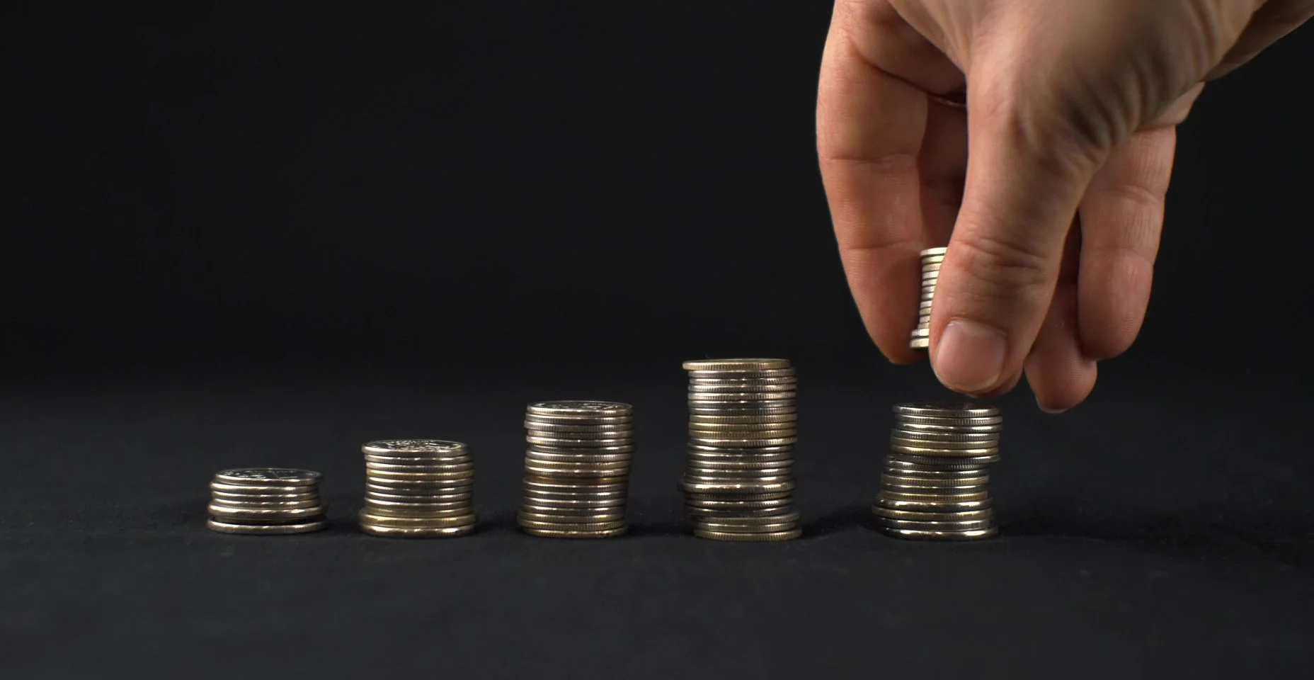 A mão relata a última pilha de moedas em um fundo preto. Cinco pilhas de moedas estão em uma fileira e o homem relata a última pilha de moedas. Aumentando o conceito de finanças.
