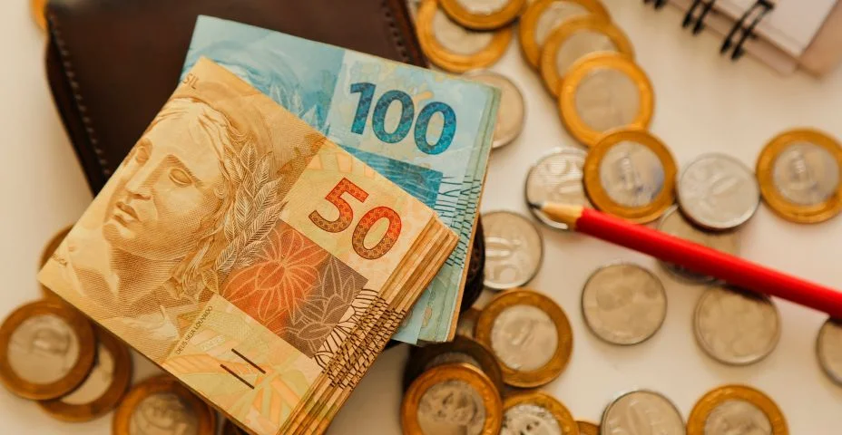 Cinquenta notas reais e outras notas e moedas sobre a mesa com carteira, lápis e caderno