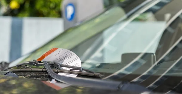 Você está diante de um carro estacionado. No vidro da frente — o para-brisa — há um papel preso sob o limpador. Esse papel parece ser uma multa de estacionamento, porque tem uma faixa laranja no topo e está cheio de texto impresso. Atrás do carro, há uma placa azul com a letra "P", que normalmente indica que é uma área de estacionamento. A cena está iluminada pela luz do dia, e ao fundo há vegetação, mas está desfocada — como se estivesse fora de foco numa fotografia.