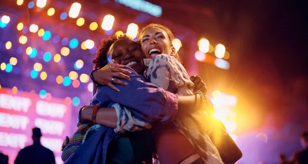 Amigas alegres abraçadas em frente ao palco da música durante o festival de verão à noite.
