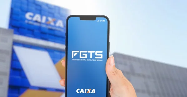 Mão feminina segurando um iPhone 13 Pro com o aplicativo do FGTS na tela. Fundo de Garantia do Tempo de Serviço (FGTS). Agência desfocada ao fundo.