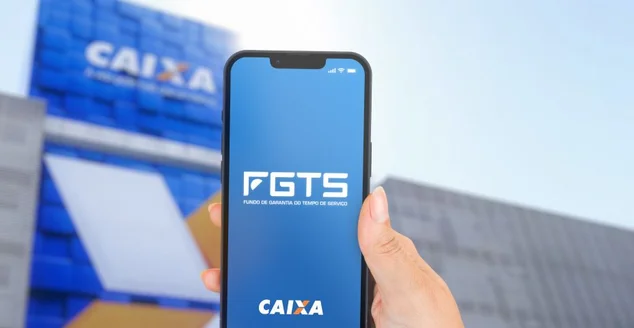 Mão feminina segurando um iPhone 13 Pro com o aplicativo do FGTS na tela. Fundo de Garantia do Tempo de Serviço (FGTS). Agência desfocada ao fundo.