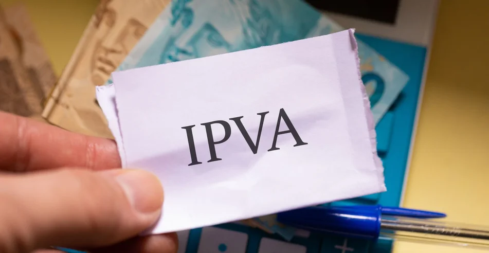 A sigla IPVA referente ao Imposto sobre a Propriedade de Veículos Automotores escrita em um pedaço de papel que um homem está segurando. Notas do Real Brasileiro e uma calculadora.