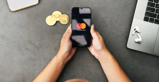 Menina segurando um smartphone com aplicativo de sistema de pagamentos Mastercard na tela na mesa de cimento com moedas de criptomoedas e computador. Ambiente de escritório.