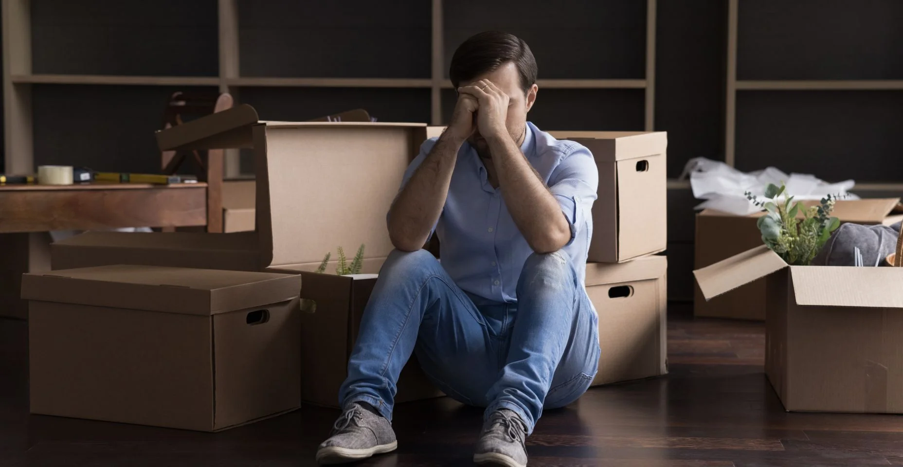 pessoa sentado no chão com varias caixas de papelões ao redor, a pessoa esta com as mãos no rosto parecendo triste