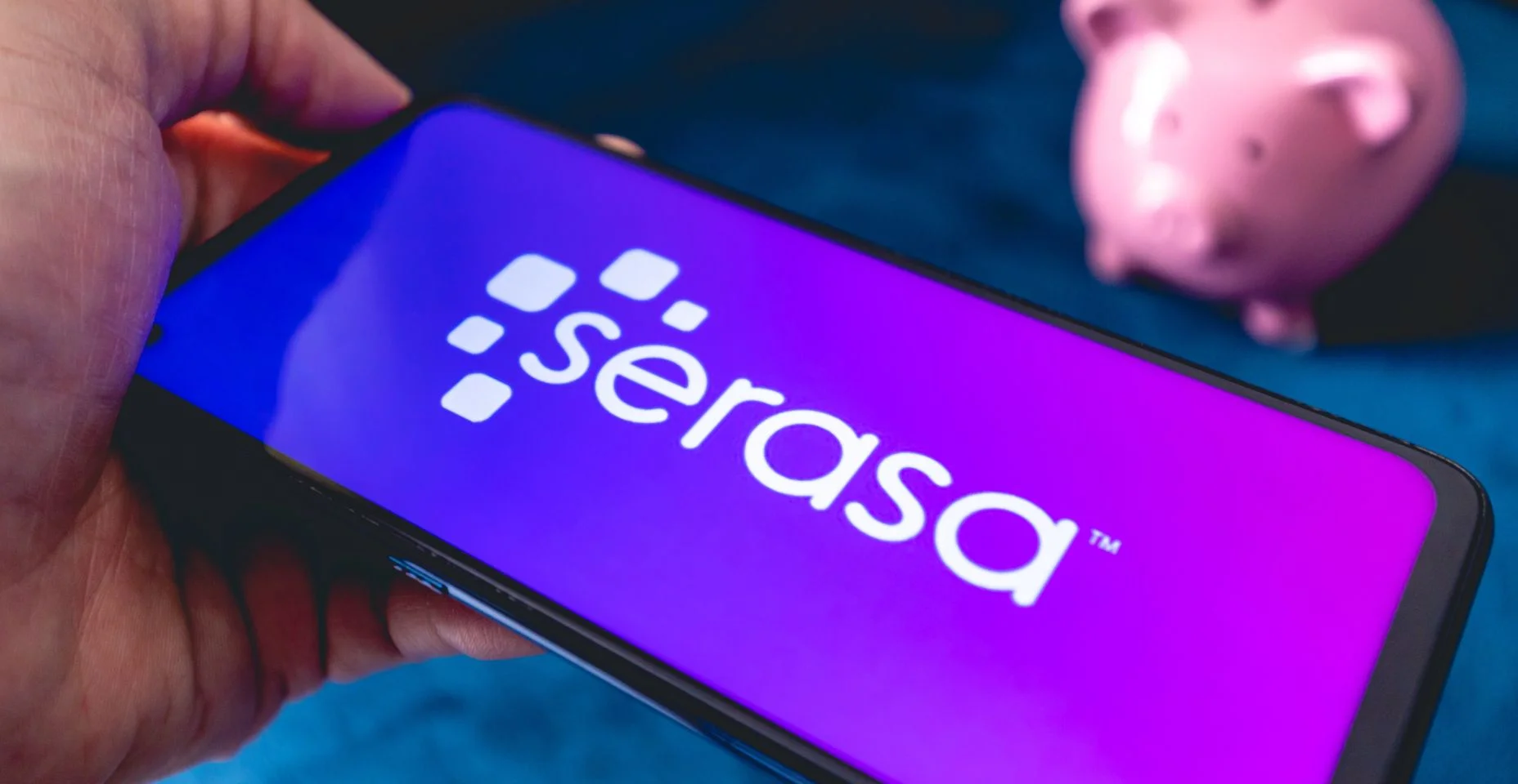 Um homem segurando um telefone celular com a logo da empresa brasileira Serasa. Um cofrinho na composição.