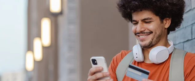 Estudante jovem olhando para o celular sorrindo, com o cartão de crédito na outra mão