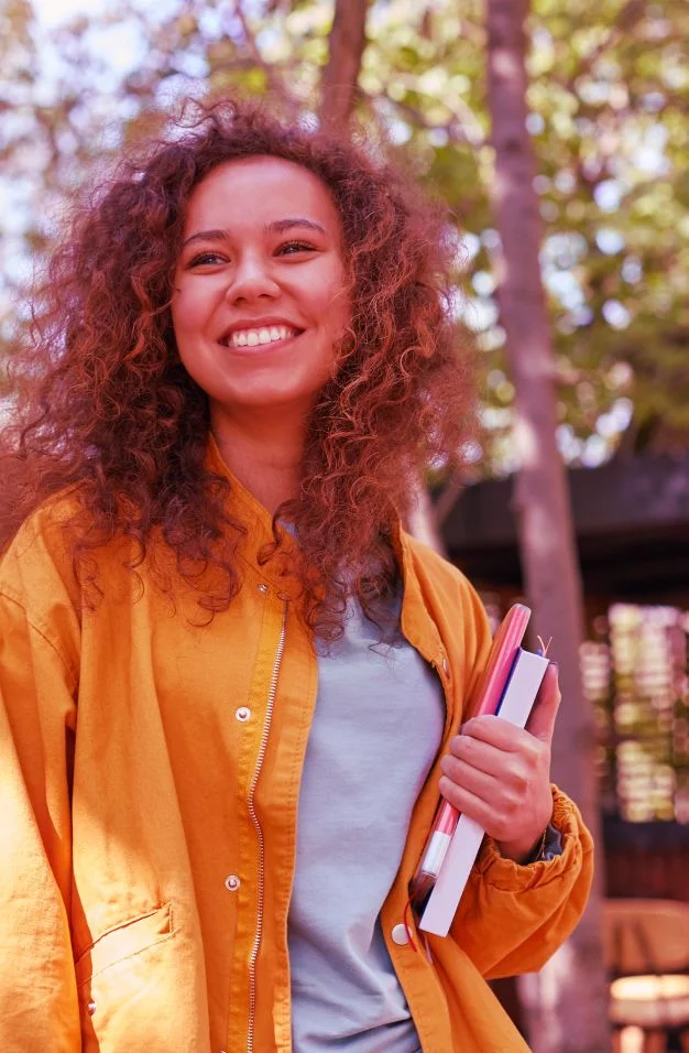 Mulher branca com o cabelo cacheado, segurando livros e está em um local aberto com árvores ao fundo