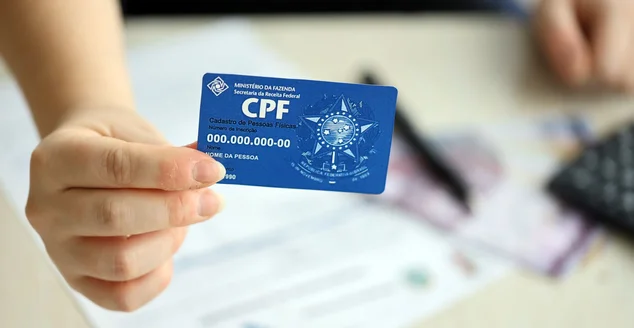 Mulher mexendo em seu notebook com o seu documento de CPF em mãos e vendo para que serve