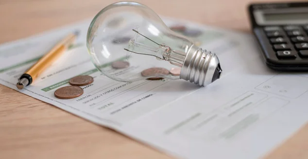 Conta de eletricidade com lâmpada, diversas moedas, calculadora e caneta em cima da mesa.