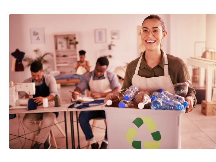 Costureira alegre segurando um balde de garrafas plásticas recicladas. Designer mestiço segurando lixo de garrafas plásticas renováveis. Jovem alfaiate reciclando garrafas plásticas de seu estúdio