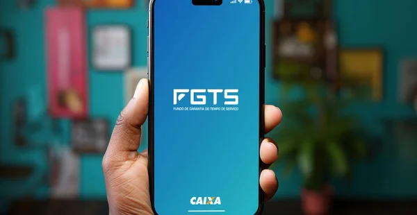 Mão negra segurando um smartphone iPhone com o aplicativo Caixa FGTS (Fundo de Garantia do Trabalhador Brasileiro) na tela. Fundo branco.