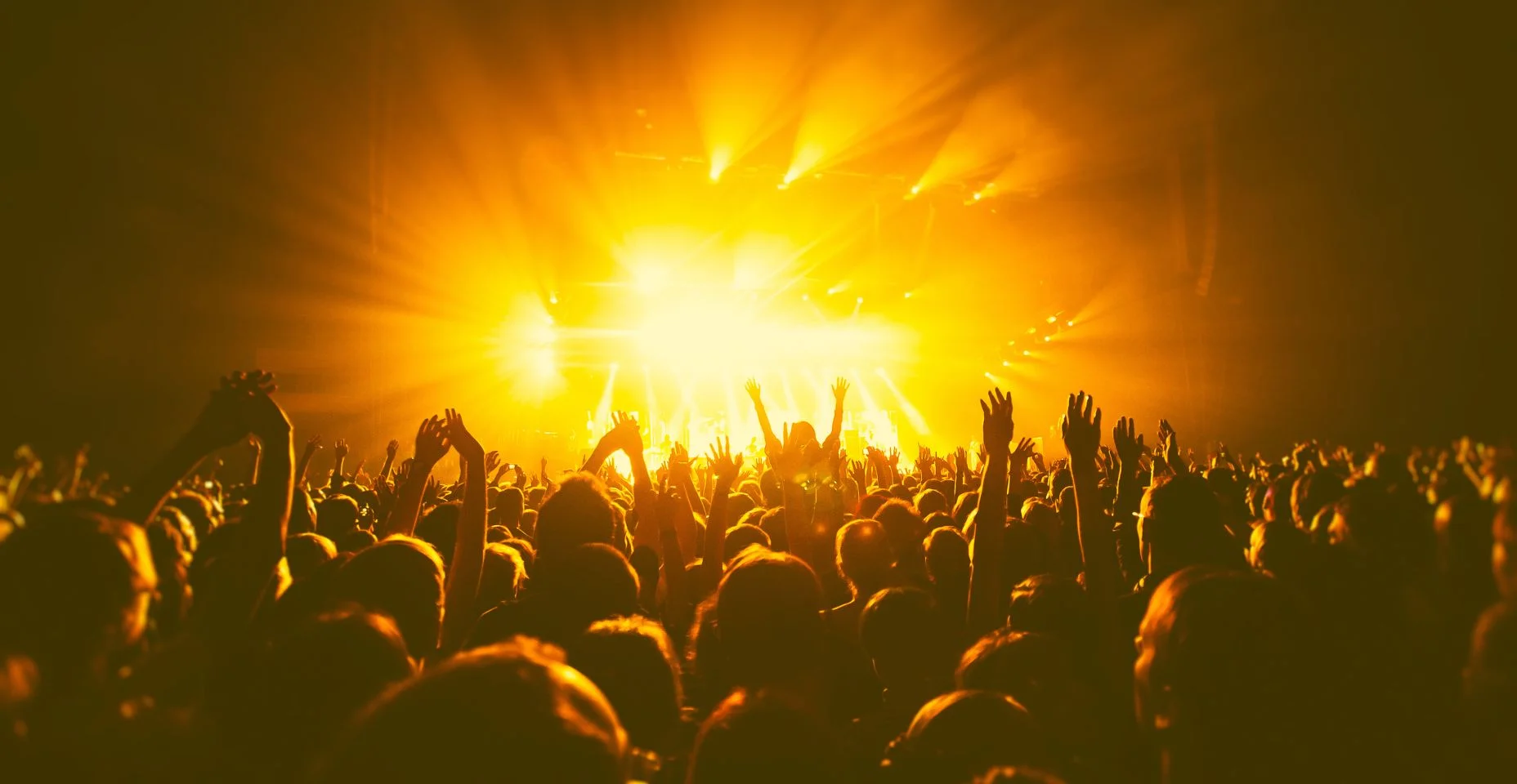 Uma sala de concertos lotada com palco de cenas laranjas e amarelas, performance de show de rock, com silhueta de pessoas, explosão de confetes coloridos disparados no ar da pista de dança durante um festival de concertos