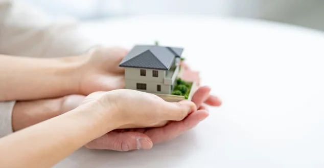 Duas mãos segurando um modelo em miniatura de uma casa, simbolizando proteção, lar ou investimento imobiliário.
