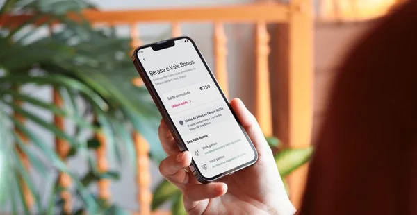 Pessoa segurando um smartphone com a tela exibindo informações do Vale Bônus da Serasa, incluindo saldo acumulado de R$170 e opções como pagar pessoas, boletos e ver extrato. Ao fundo, há vegetação desfocada e elementos de madeira.