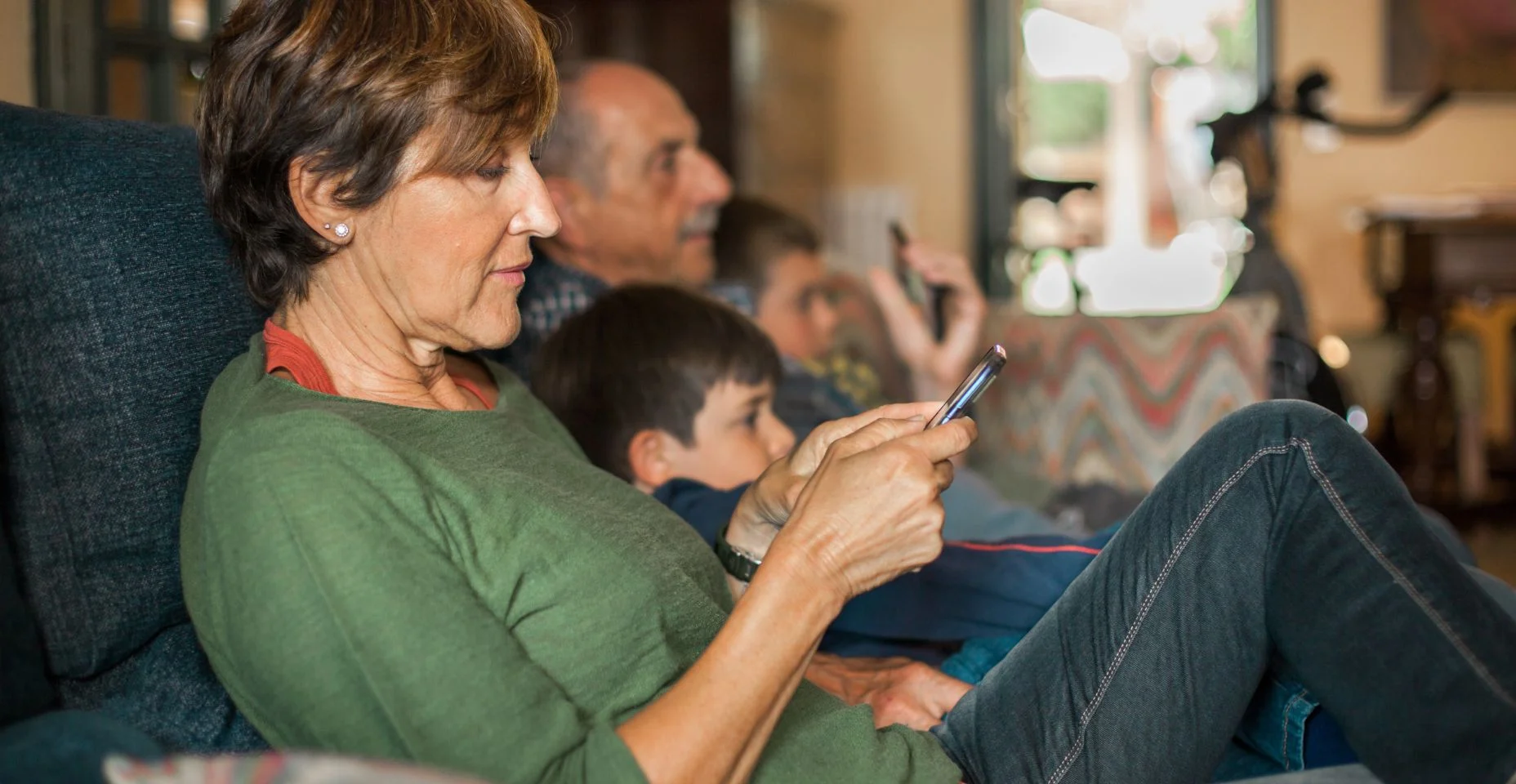 Avó sentada no sofá olha o celular enquanto a família assiste televisão.