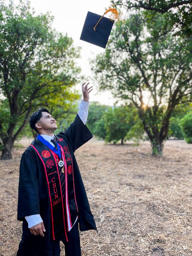 Raphael Zambrano standing outdoors in his graduation robe throwing his graduation cap in the air.