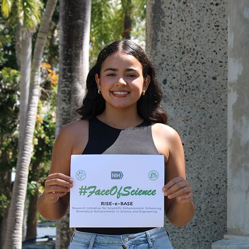 Karina Ruiz Rivera holding a sign with #FaceOfScience RISE e-BASE written on it.