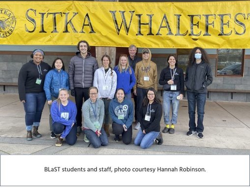 BLaST students and staff standing outside, under a large, yellow Sitka Whalefest banner in Sitka, Alaska.