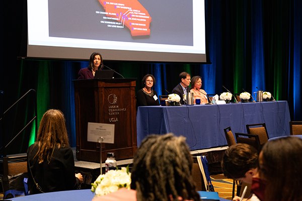 Dorota Huizinga, PhD, stands at the podium presenting while the other three panelists are seated at a table.