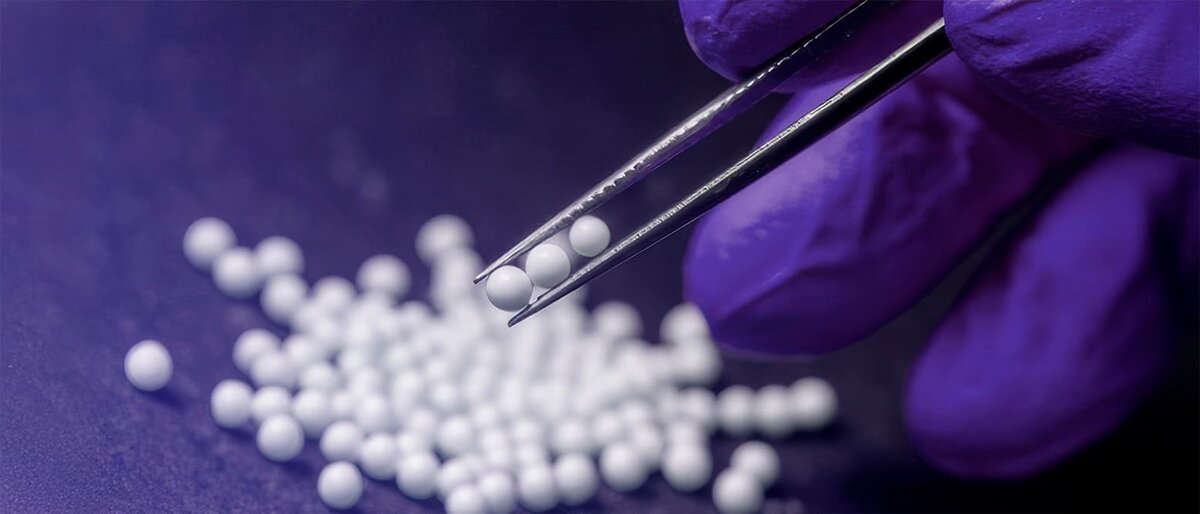 Close-up of a gloved hand holding lyophilized beads with tweezers, with more beads scattered on a dark surface in the background