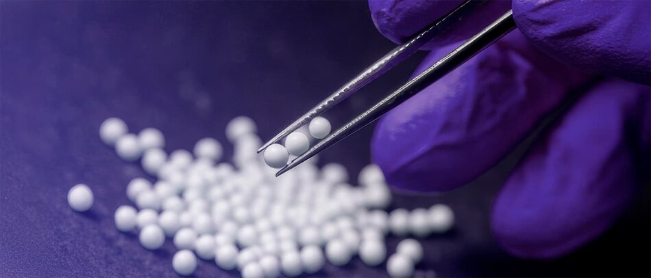 Gloved hand using tweezers to pick up small white beads on a lab surface.