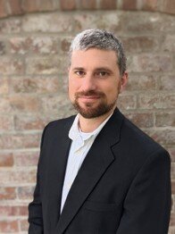 A man with short gray and dark hair, a beard, and a black suit jacket poses against a brick wall background.