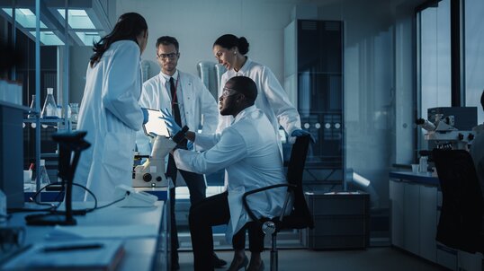 A group of four scientists - three sitting and one standing - huddle around a tablet.