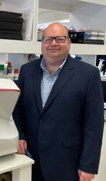 Dr. Mike Spencer in a navy suit and glasses smiling in a lab setting