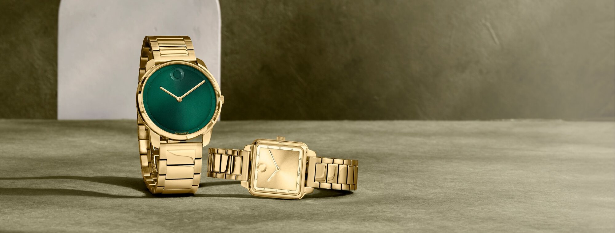 Movado Trend men’s green-dial gold watch beside women’s gold watch with shaped dial