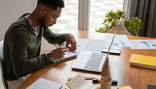 person writing a notebook at a desk, in front of an open laptop