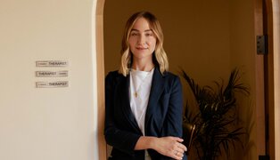 woman standing and leaning against a doorway, signs saying "therapist in session" or "therapist available" are visible to the left