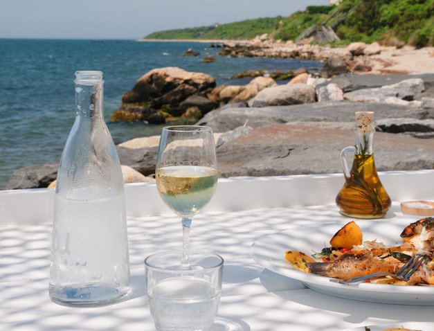 Glass of white wine alongside Sea Bass Entree with water view of Fort Pond Bay