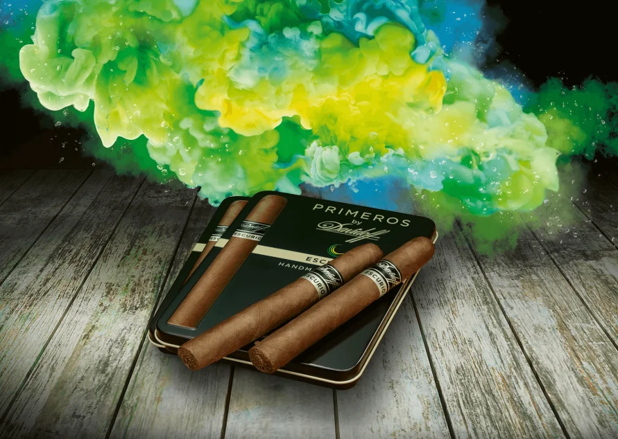 Eine Ausstellung von Primero-Zigarren, angeordnet neben einer eleganten Dose, mit lebhaftem, farbenfrohem Rauch, der im Hintergrund auf einer Holzoberfläche wirbelt.