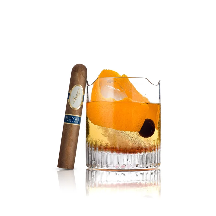 Cigar beside a whiskey glass with orange peel and cherry on a white background.