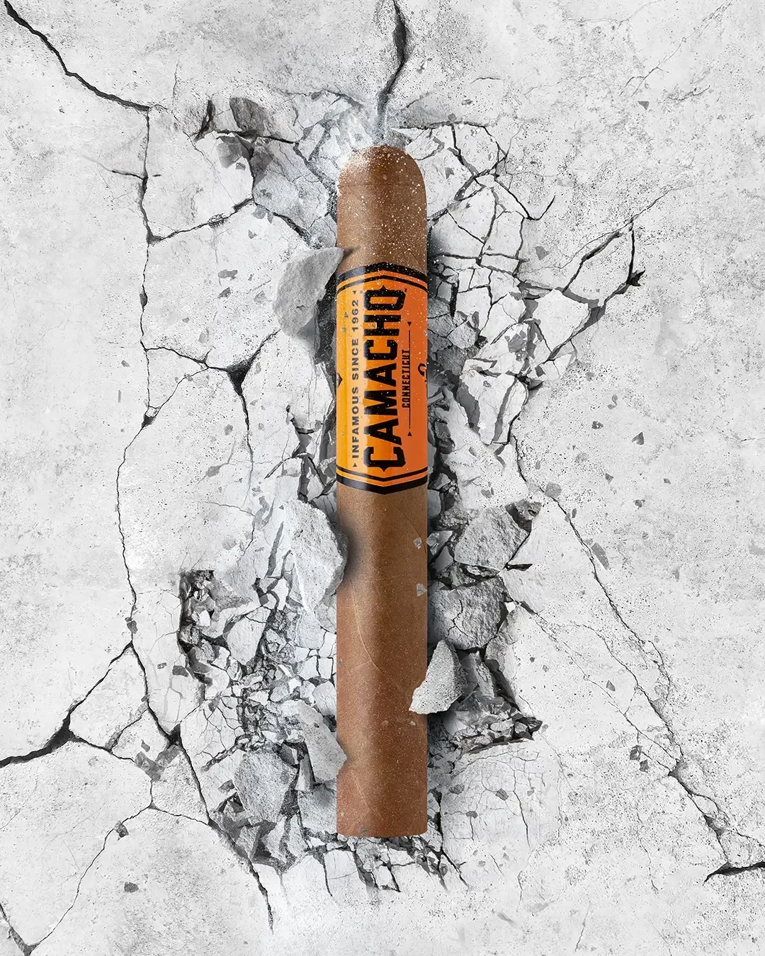 Eine Camacho-Zigarre ragt aus rissigem Beton hervor mit ihrer markanten orangefarbenen Banderolle.