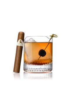 Bernsteinfarbener Cocktail mit Kirschgarnitur neben einer Zigarre auf reflektierender Oberfläche.