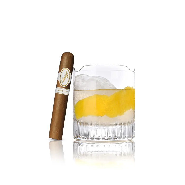 Davidoff Grand Cru cigar beside a whiskey glass with lemon twist and ice on a reflective surface.